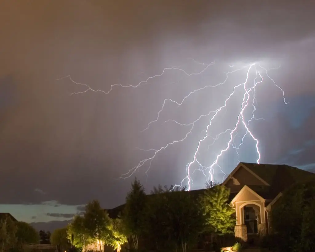 A vivid lightning storm illuminates the night sky over a suburban neighborhood. Branches of lightning stretch across dark clouds, casting a dramatic glow on houses and trees below. The scene captures the power and beauty of nature.