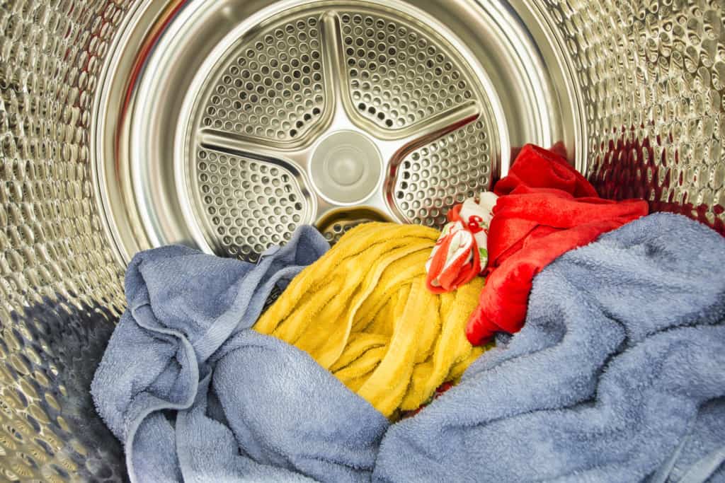 An open washing machine drum containing a few colorful towels: one yellow, one blue, and one red. The interior of the drum is metallic with a circular, perforated pattern, appearing clean and shiny.