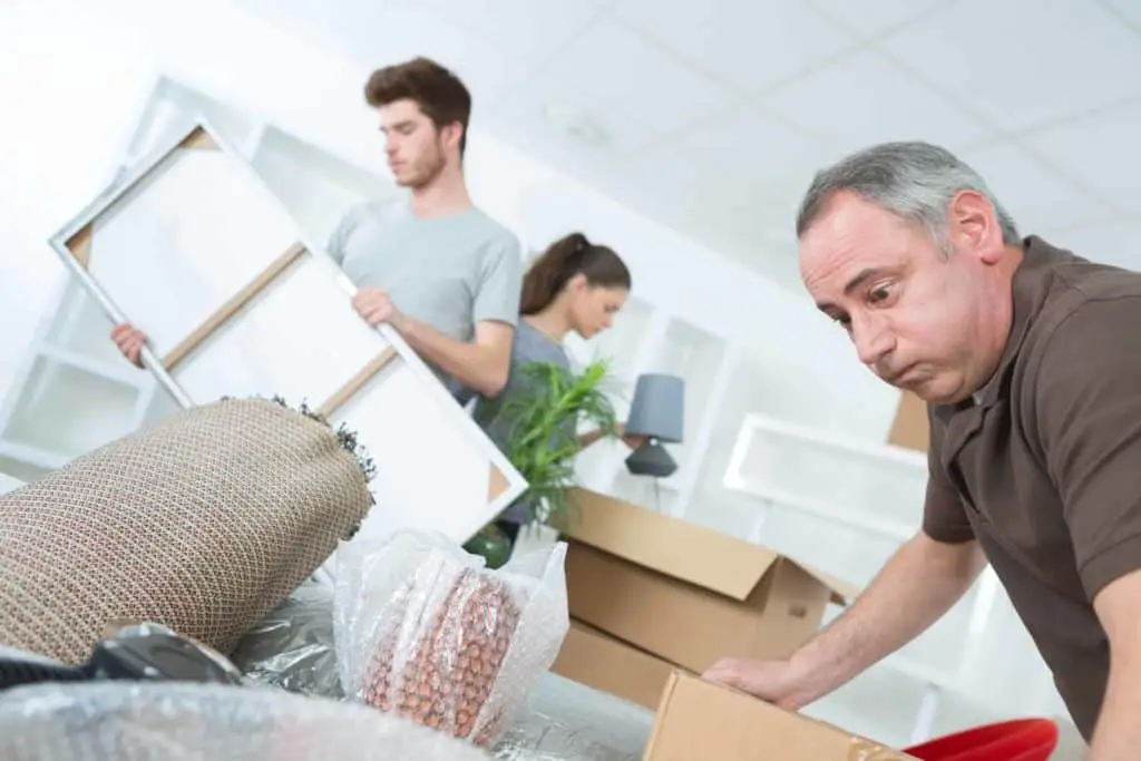 Three people are busy unpacking or packing various household items into cardboard boxes in a bright room. In the foreground, a man is focused on his task, while another man and a woman can be seen in the background handling picture frames and other items.