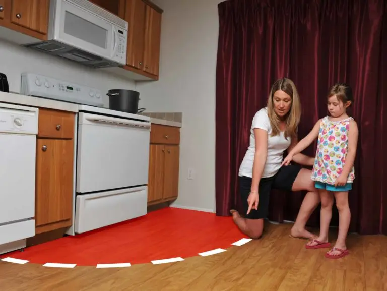A woman kneels on the kitchen floor pointing at a red safety zone marked with white dashes around the stove. A young girl stands next to her, looking at the red area. The kitchen includes a stove, oven, microwave, and wooden cabinets.