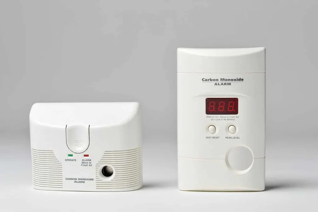 Two different styles of carbon monoxide detectors are placed side by side. The left device is compact with a test/reset button, while the right device is taller with a digital display showing "8.8.8" along with buttons labeled "Test/Reset" and "Peak Level.
