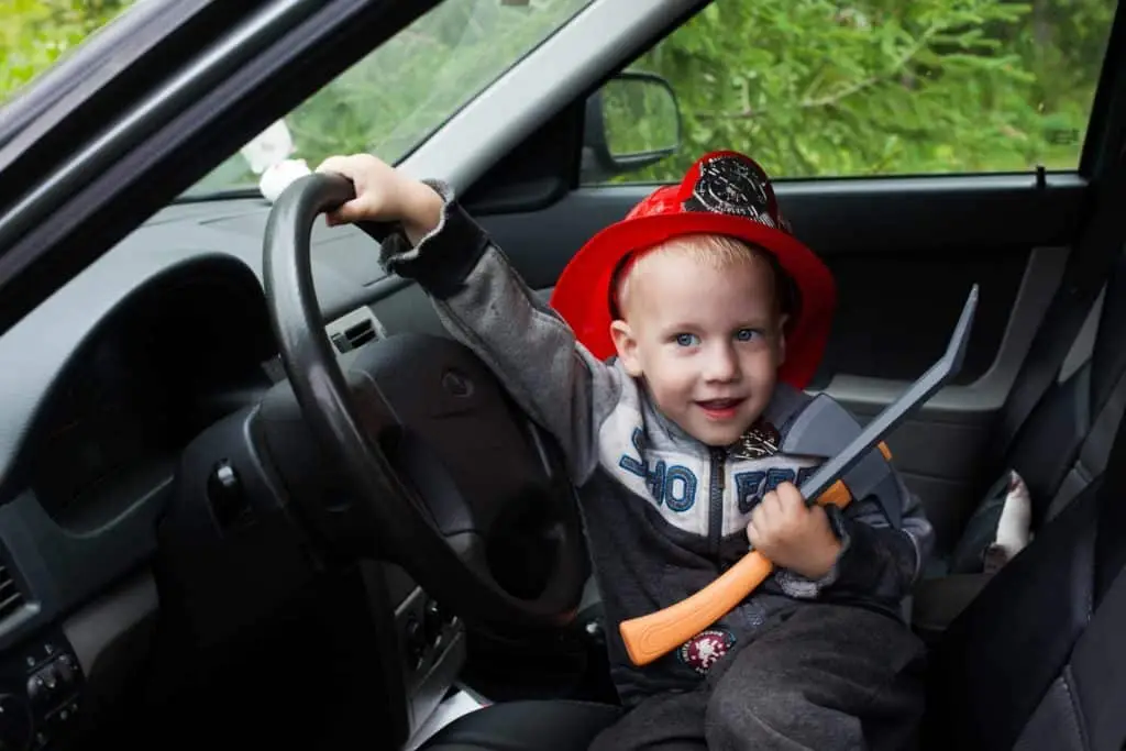 A young child wearing a red firefighter hat sits in the driver's seat of a car, holding the steering wheel with one hand and a toy axe in the other, while smiling at the camera. Green trees are visible through the car window in the background.