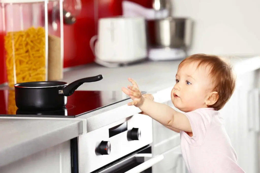 A baby in a white onesie reaches up towards a black saucepan on a stove in a modern kitchen. The baby is on tiptoes, extending both arms towards the pan. The background includes kitchen appliances and jars of pasta.
