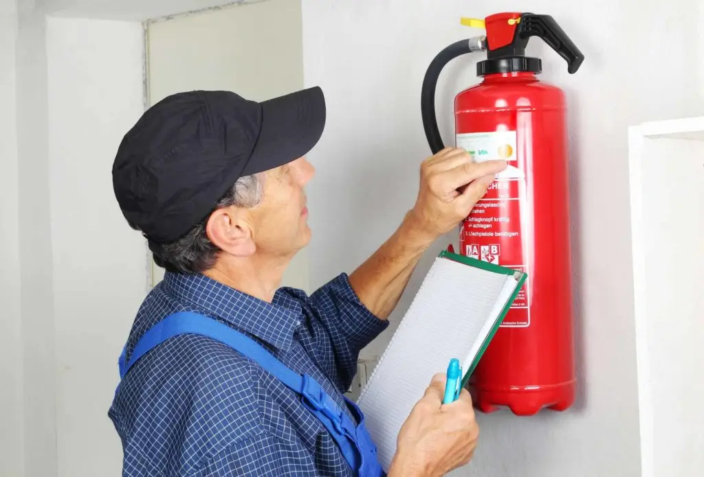 A man in a black cap and blue overalls is inspecting a red fire extinguisher mounted on a wall. He is reading the label on the extinguisher while holding a clipboard and pen in his hands. The room appears to be indoors and has white walls.