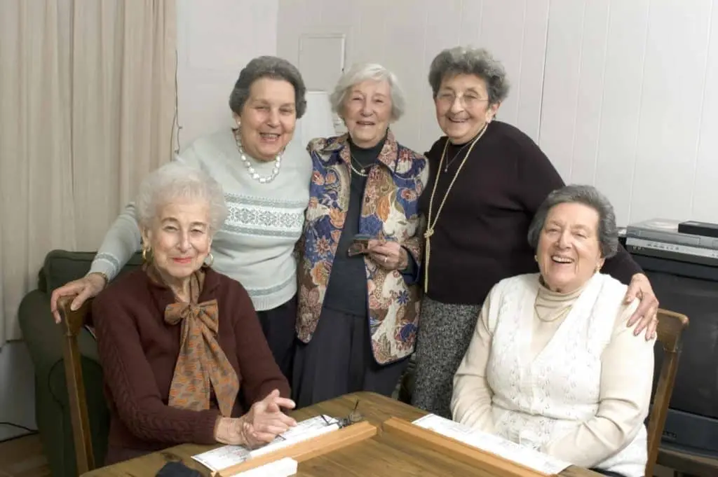 Five elderly women are gathered together, with three standing at the back and two sitting at the front. They are smiling and appear to be enjoying each other's company. In front of the seated women, there is a table with a game or activity laid out on it.