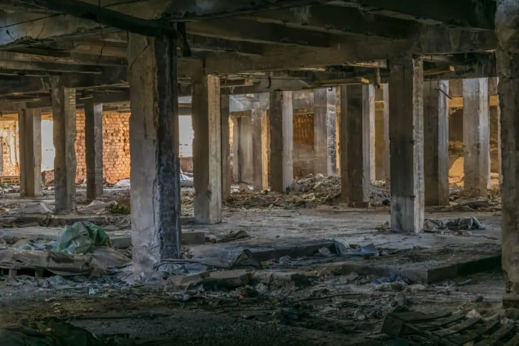 A dilapidated, abandoned building interior with multiple concrete pillars, crumbling walls, and scattered debris on the ground. The scene is dimly lit with sunlight filtering through broken sections of the structure, highlighting the decay.