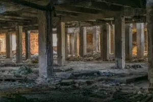 A dilapidated, abandoned building interior with multiple concrete pillars, crumbling walls, and scattered debris on the ground. The scene is dimly lit with sunlight filtering through broken sections of the structure, highlighting the decay.