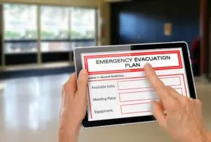 A person holding a tablet displaying an "Emergency Evacuation Plan." The screen shows sections for "Available Exits," "Meeting Place," and "Equipment," with blank fields. The background is a blurred indoor setting, appearing to be a public or office space.