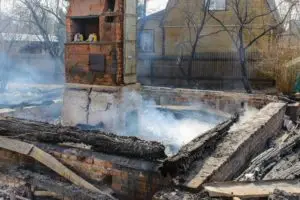 Remains of a brick house with charred wooden beams and smoke rising from the debris. A partially intact brick chimney stands in the center. In the background, leafless trees and wooden fences are visible, suggesting recent fire damage.