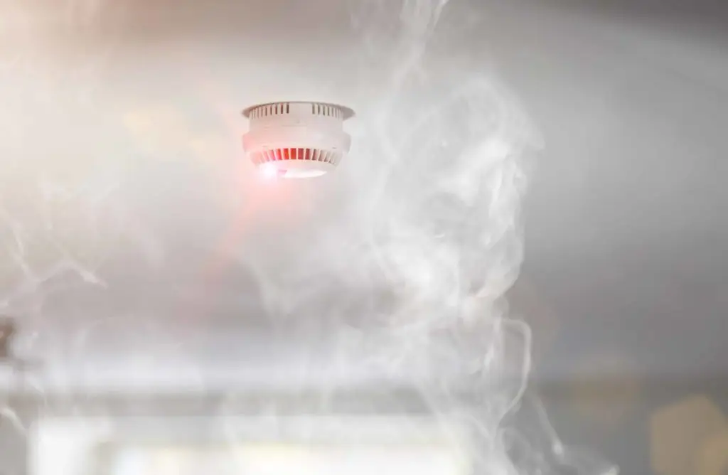 A smoke alarm with a red indicator light is mounted on a ceiling. Smoke surrounds the alarm, suggesting it has been activated due to the presence of smoke. The background is blurred, focusing attention on the alarm and the smoke.