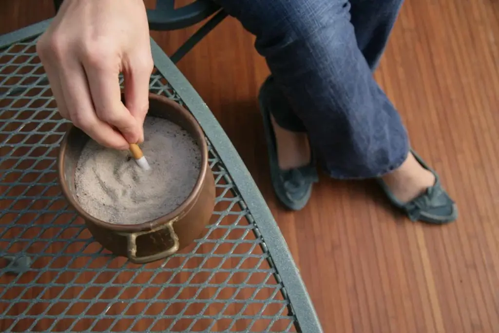 A person sits on a chair with legs crossed, stirring a hot beverage in a brown mug using a cigarette. The mug is placed on a metal mesh table, and the person is wearing blue jeans and gray shoes. The background is a wood floor.