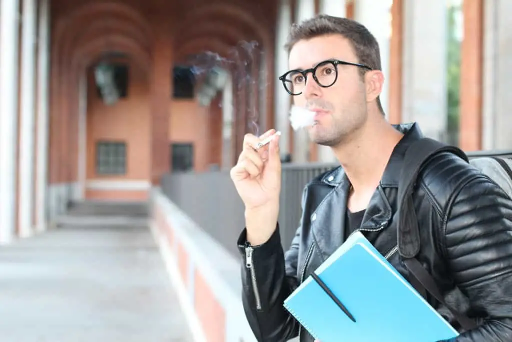 A man wearing glasses and a black leather jacket is smoking a cigarette while holding a blue notebook. He stands in an outdoor hallway with arched ceilings and red brick walls in the background.
