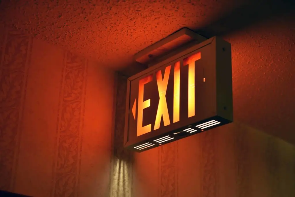 A glowing red and orange exit sign with an arrow pointing left, mounted on the ceiling of a room with textured wallpaper. The sign casts a warm light on the surrounding area, indicating the direction to the exit.