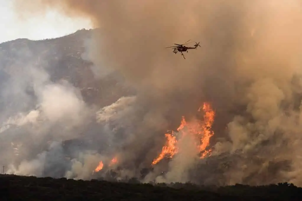 A helicopter flies over a wildfire on a mountainside engulfed in flames and thick smoke. The fire has spread widely, with large orange flames and dense clouds of gray smoke rising into the sky. The lower part of the image shows dark green vegetation.