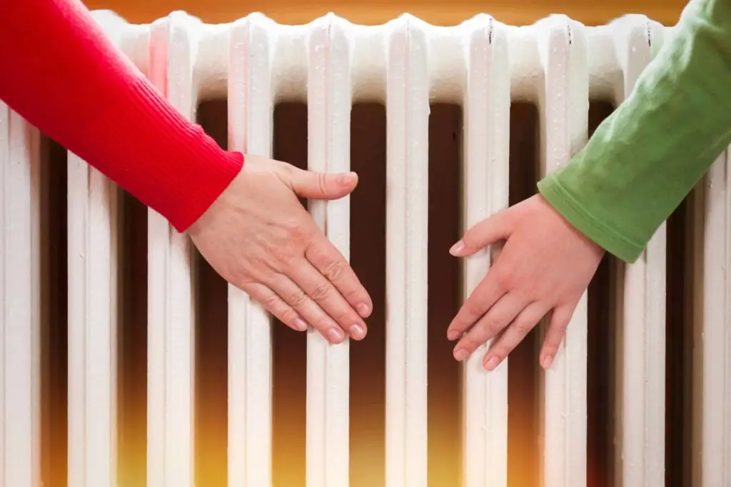Two hands, one in a red sleeve and the other in a green sleeve, are placed near a white radiator or heater. The image suggests the concept of staying warm or heating during colder weather. The background is softly out of focus with a warm tone.