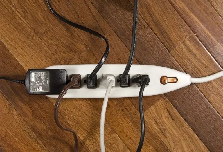 A white power strip with six outlets lies on a wooden floor. Four of the outlets are in use, with various black, white, and brown cords plugged in. One plug is an AC adapter, while the other three are standard power plugs. The remaining two outlets are empty.