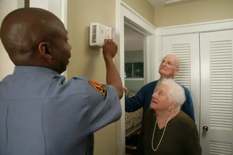 A firefighter in uniform is installing or checking a smoke detector in a home. An elderly couple stands nearby, watching attentively. The man is wearing a blue shirt and the woman is wearing a dark green sweater with a necklace. They are in a well-lit hallway.