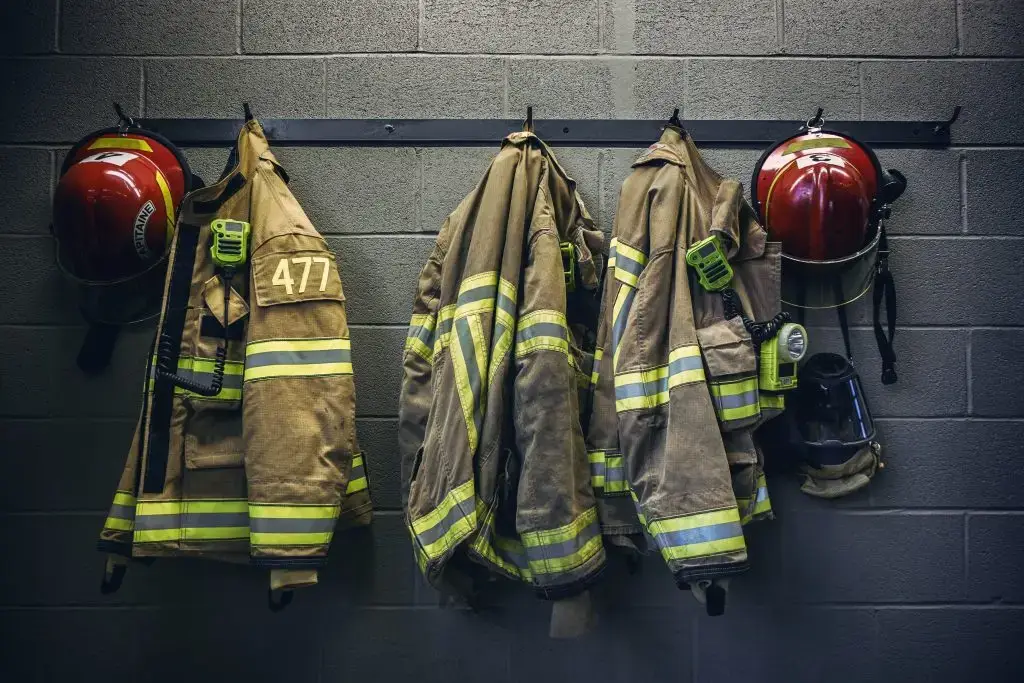 Three firefighter jackets and two helmets hanging on a wall hook. The jackets have reflective stripes and one is labeled "477". A firefighter mask is also hanging on the right side. The wall is made of gray brick.