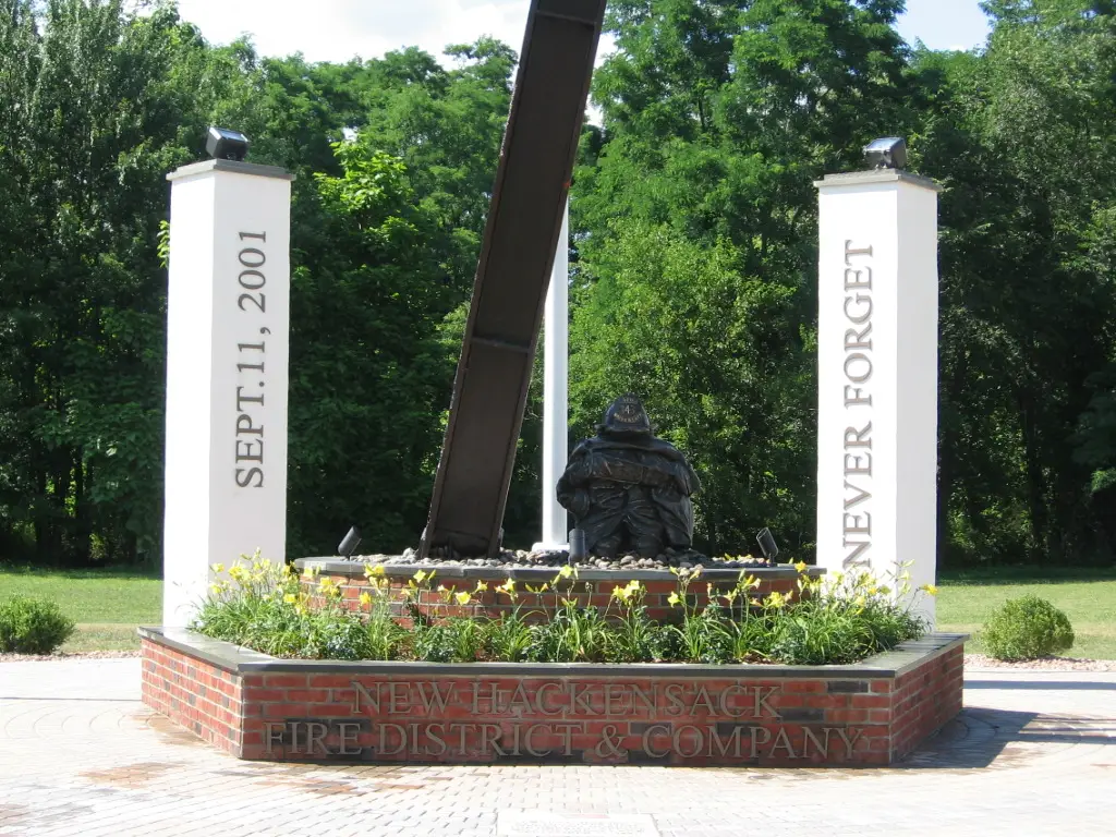 A memorial featuring a section of a steel beam from the World Trade Center attacks. The beam is flanked by two white columns reading "SEPT. 11, 2001" and "NEVER FORGET." A firefighter statue and flowers are also present. The base reads "NEW HACKENSACK FIRE DISTRICT & COMPANY.