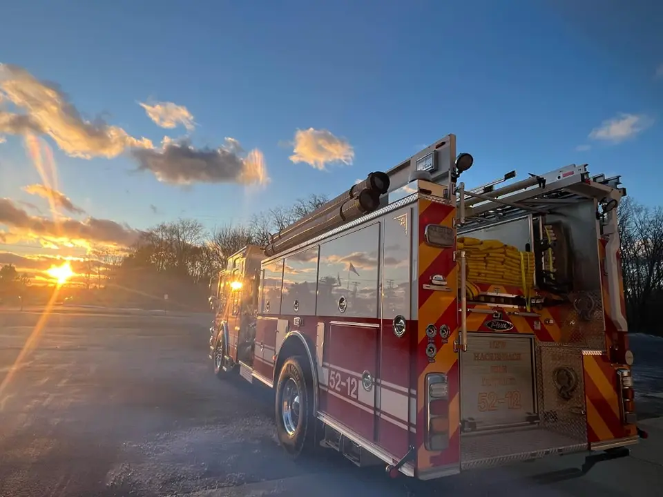 A fire truck with red and yellow markings is parked on a road at sunset. The sun is low in the sky, casting a golden light and long shadows. Bare trees are visible in the background under a partly cloudy sky.