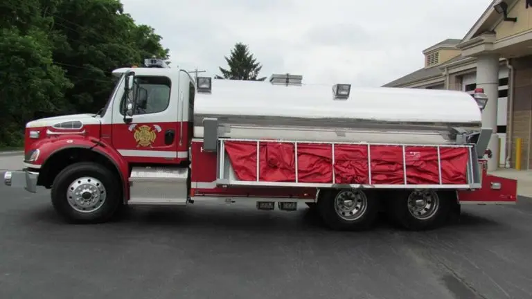 A red and white fire truck with a water tanker is parked outdoors near a building. The truck features several compartments and equipment storage areas on its side. The background shows trees and part of the building.