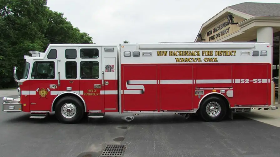 A red and white fire truck labeled "New Hackensack Fire District" and "Rescue One" is parked outside a building with a sign that reads "New Hackensack Fire District." The truck has decals and emergency lights, and the building has columns at the entrance.