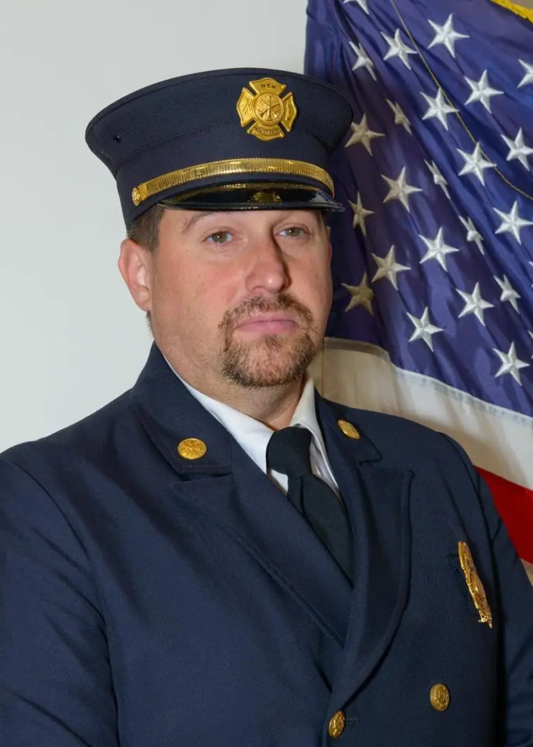 A uniformed firefighter stands in front of an American flag. He has a trimmed beard and mustache, wearing a dark blue double-breasted coat with brass buttons and a matching cap adorned with a fire department badge. He has a serious expression.