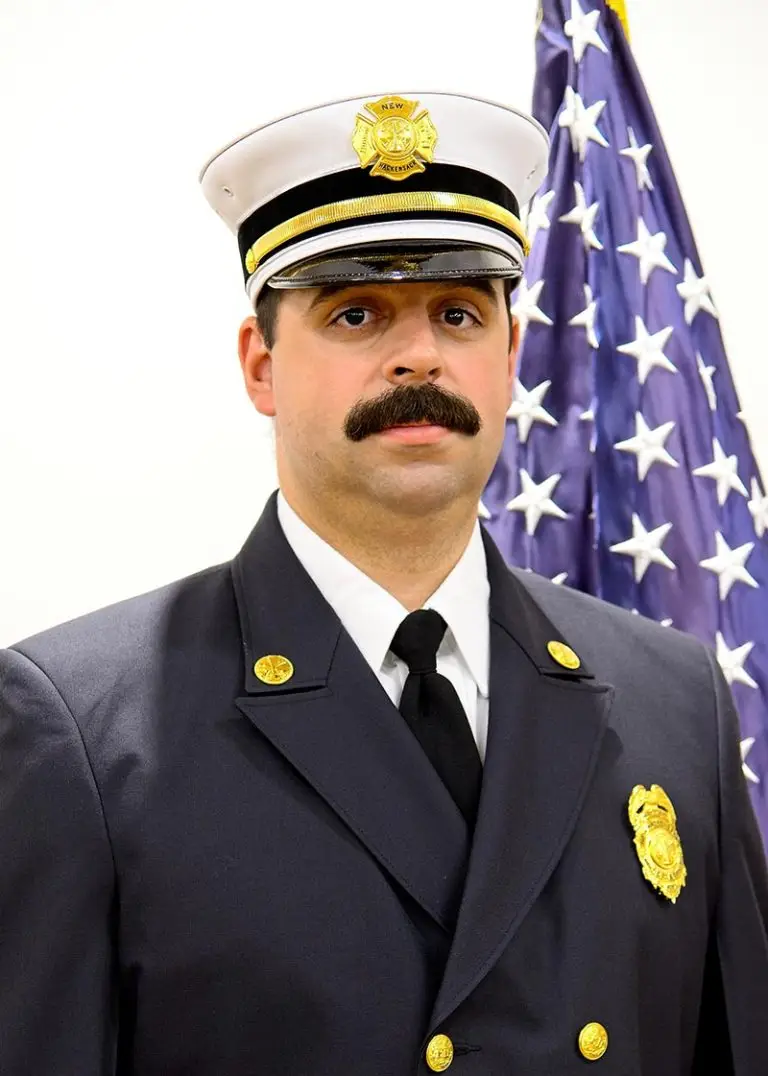 A man in a formal fire department uniform stands in front of a U.S. flag. He wears a white peaked cap with an emblem, a dark jacket adorned with badges, and a black tie. He has a mustache and looks directly at the camera.