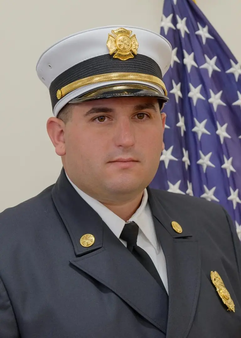 A man wearing a formal dark uniform and a white hat with a gold badge stands in front of an American flag. He has a serious expression on his face and is adorned with several gold buttons and insignia on his uniform.