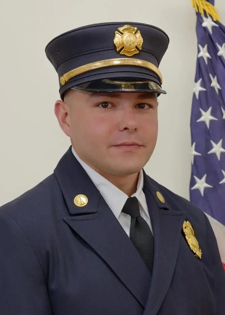A man dressed in a firefighter's formal blue uniform, complete with a peaked cap adorned with a gold badge, stands beside an American flag. He has a composed expression and is facing the camera.