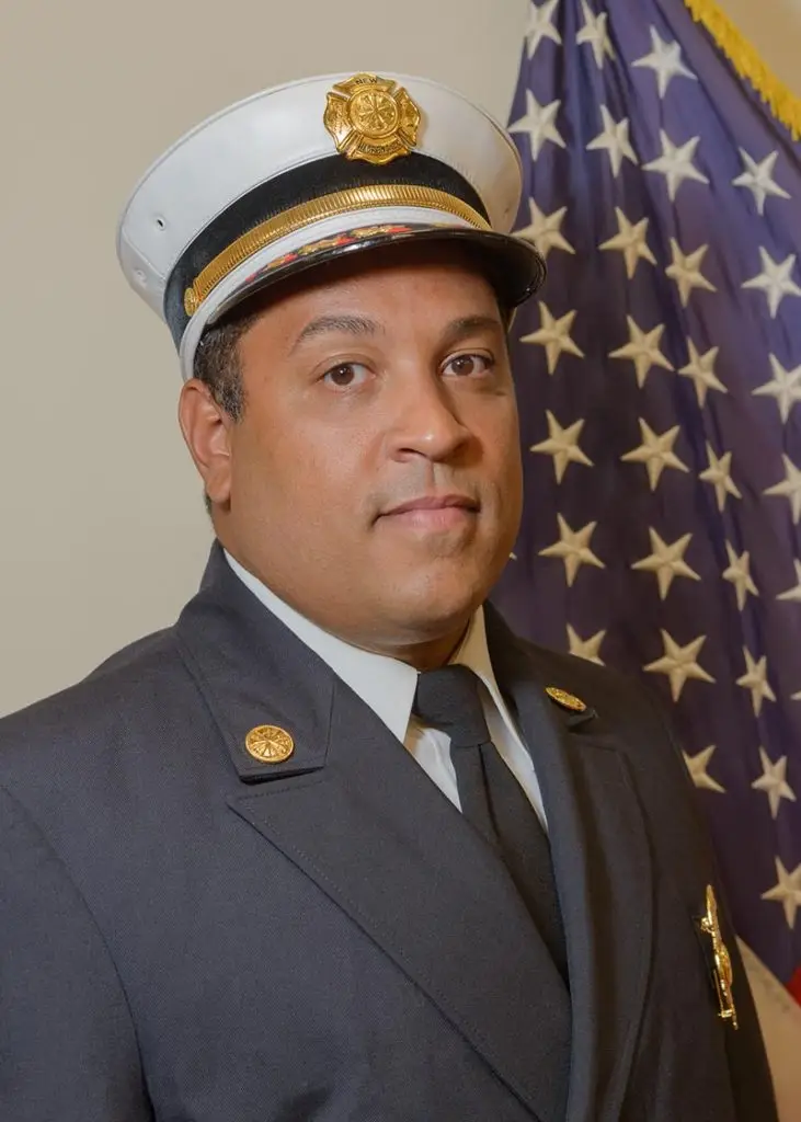 A man dressed in a formal firefighter's uniform, including a white cap with a badge, poses near an American flag. He has a serious expression and stands against a plain background.