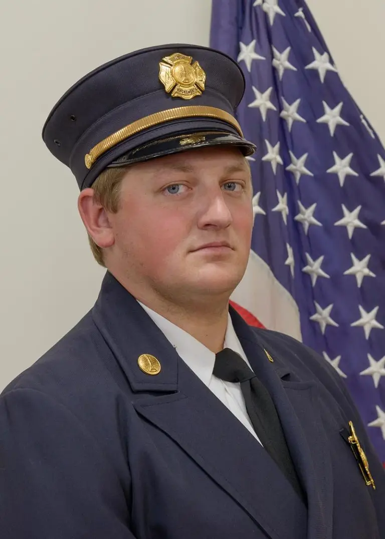 A man in a navy blue firefighter dress uniform stands in front of an American flag. He is wearing a hat with a gold badge, has a serious expression, and is looking directly at the camera.