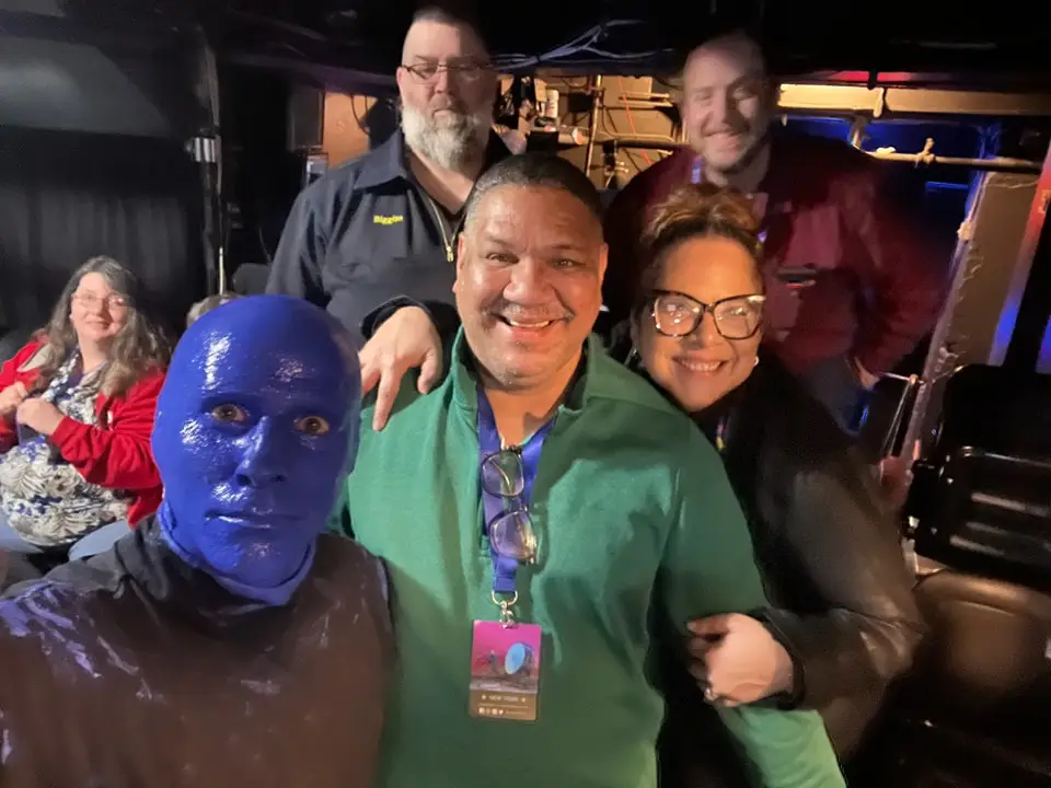 A group of five people smiling for the camera indoors. The person on the left is painted blue and wearing a black outfit. The others are casually dressed; three men and one woman are present. The person in front is wearing a blue lanyard with a badge.