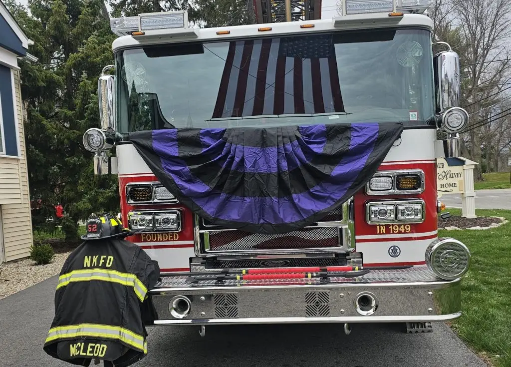 A firetruck adorned with black and purple bunting has a firefighter in full gear kneeling in front of it. An American flag is visible through the windshield. The firetruck displays "NKFD" and "52" emblems, and the building nearby has the sign "Valley Home.