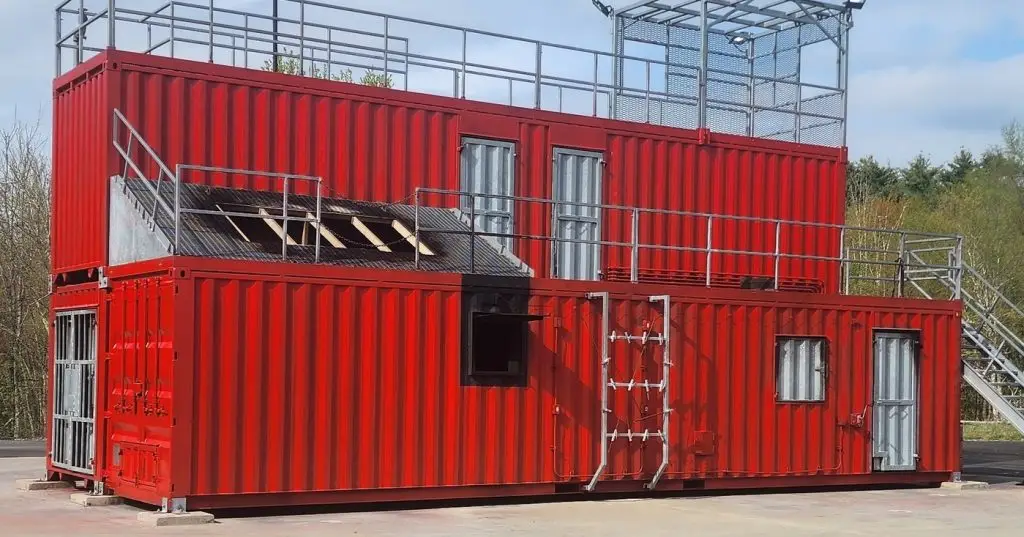 A red two-story building made from shipping containers, featuring metal railings and stairs. The structure includes windows, doors, and a mesh cage on the roof. The background shows greenery and a cloudy sky.